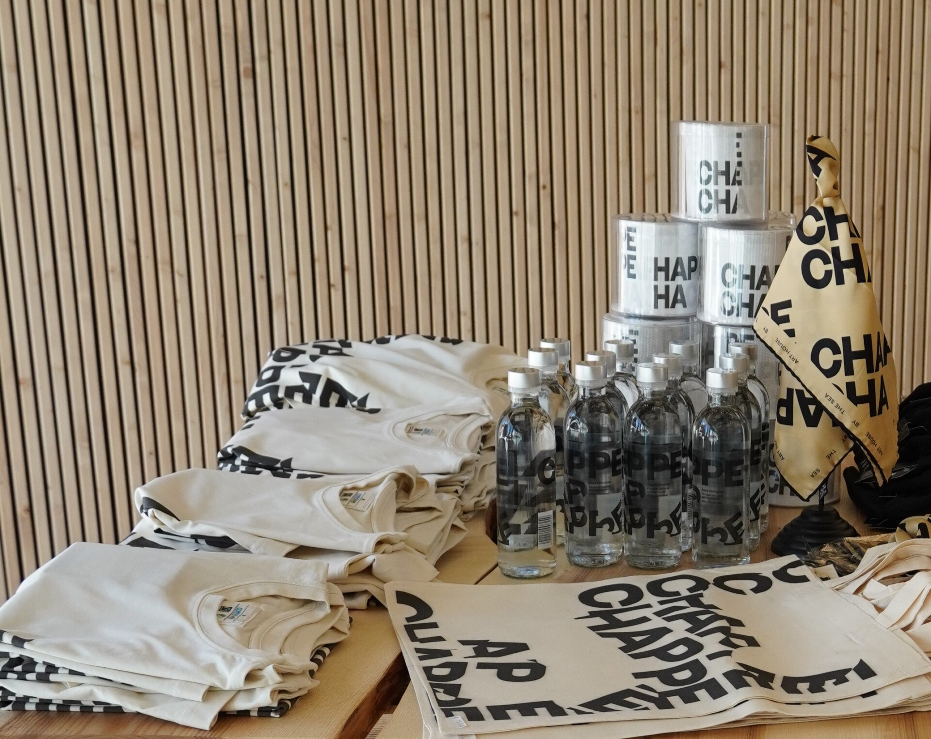 Chappe products on a table against a wooden wall. The products consist of t-shirts, water bottles, canvas tote bags, toilet paper, and a scarf. All products have the Chappe logo on them.