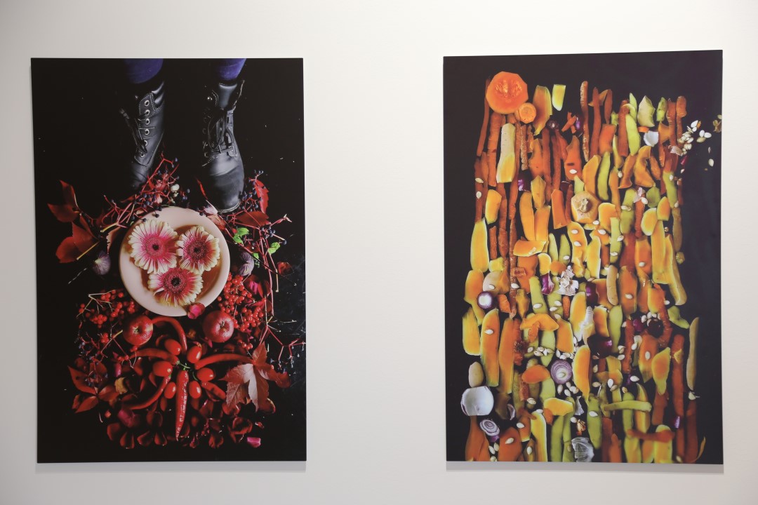 Två fotografier av Tanja Marjaana Heikkilä. Fotogarfier till höger visar fötter klädda svarta skor vid en vit tallrik med tre blommor, omrigat av röda frukter och växter. Växterna är placerade i ett mönster runt tallriken. Fotografier till höger föreställer en yta som består av rotfruktsskal, lökskal och annat organiskt marerial.